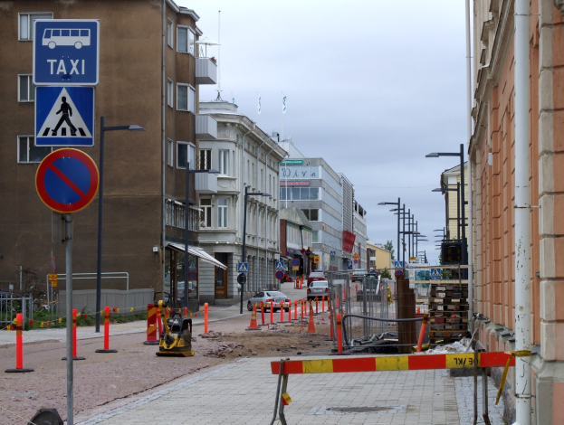 Eine Stadtstraße mit Gebäuden, Straßenlaternen, Schildern, Verkehrskegeln, Fahrzeugen, Absperrpoller, Bäumen und einer Baustelle mit Verkehrszeichen unter einem bewölkten Himmel.