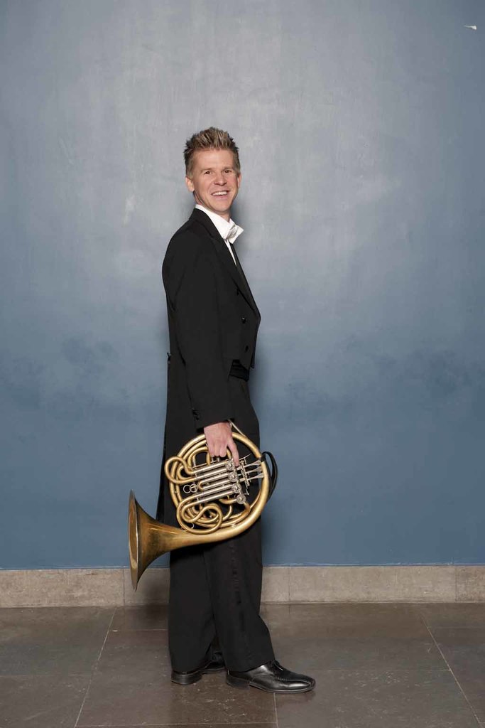 Ein Mann steht auf einem Boden, lächelt und hält ein French Horn in der Hand, mit einer Wand im Hintergrund.