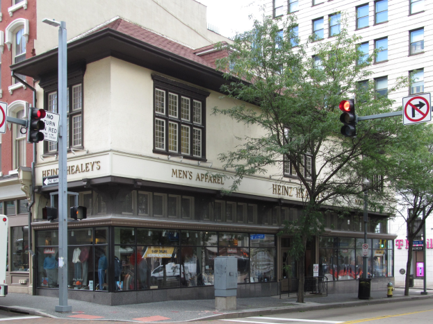 Eine belebte Straßenkreuzung mit einem geparkten Bus, Gebäuden, Bäumen, Laternen, Verkehrsampeln, Schildern und einem Kaufhaus mit Schaufensterpuppen im Hintergrund unter einem sichtbaren Himmel.