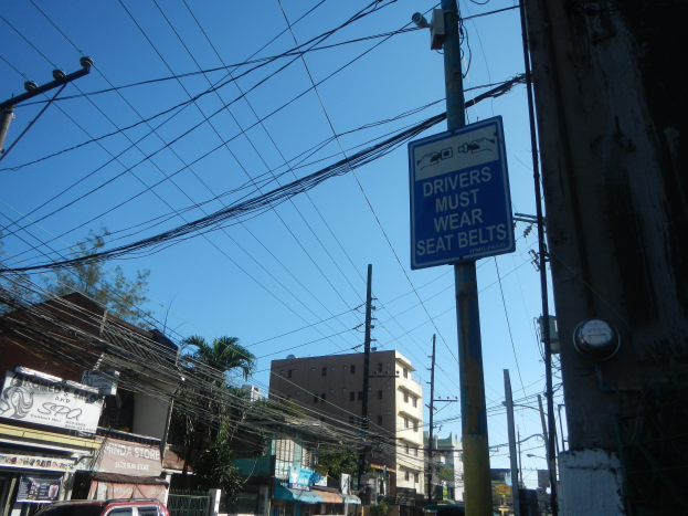 Stadtstraße mit fahrenden Autos, Strommasten mit Überlandleitungen, Gebäude mit Fenstern, Bäume, Namensschilder und ein "Fahrer müssen Sicherheitsgurte tragen"-Schild an einem Strommasten vor einem sichtbaren Himmel.