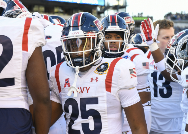 Gruppe von Männern in Football-Uniformen auf einem Feld mit einem unscharfen Publikum, Anzeigetafel mit 'Auburn vs Virginia Tech - 11/28/15 College Football Pick, Odds, and Prediction' und Himmel im Hintergrund.