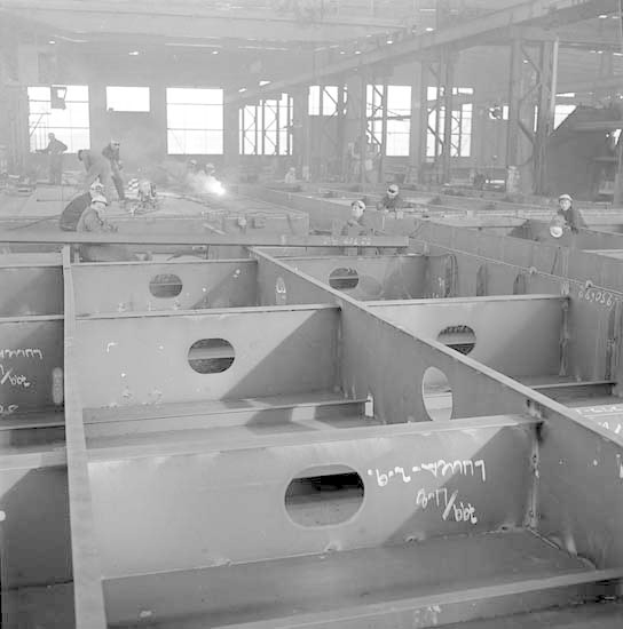 Ein Schwarz-Weiß-Foto einer Fabrik mit Arbeitern in Helmen, die an Stahlträgern arbeiten, Fenstern, Polen, Lampen und Text im Hintergrund.