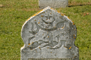 Ein steinernes Grabmal mit arabischer Inschrift, das in einem grasbewachsenen Friedhof steht.
