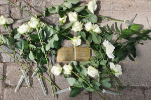 Wei├čer Rosenkranz und Kerzen mit goldenen, beschrifteten Gegenst├Ąnden vor einem Berliner Mahnmal.