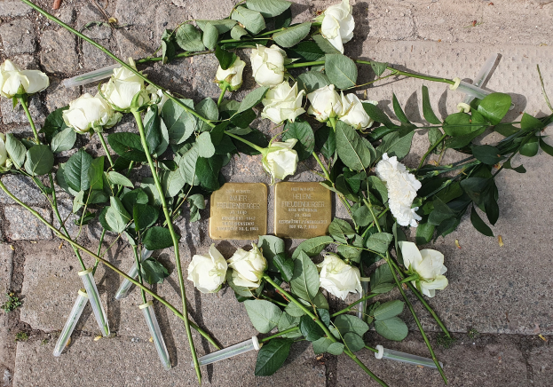 Wei├čer Rosenkranz und Kerzen mit goldenen, beschrifteten Gegenst├Ąnden vor einem Berliner Mahnmal.
