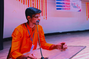 Ein Mann in einem orangen Hemd mit einem ID-Badge sitzt an einem Podium mit einem Mikrofon, das vor einem weißen Bildschirm mit einem Banner und einem schwarzen Vorhang links spricht.