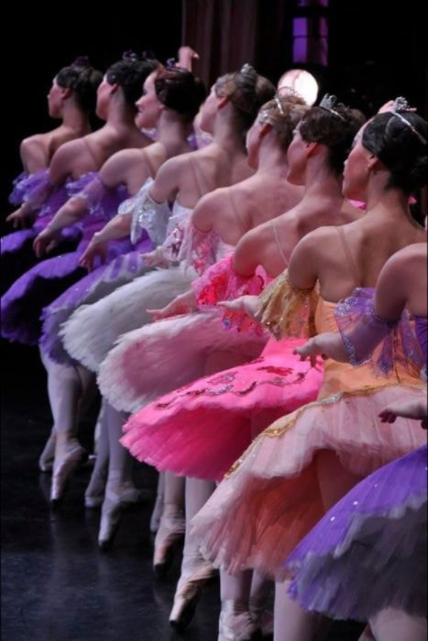 Eine Gruppe von Balletttänzerinnen in rosafarbenen Tutus sitzt auf einer Bühne, beleuchtet von einem Licht im Hintergrund, und bewegt sich in harmonischer Synchronisation.